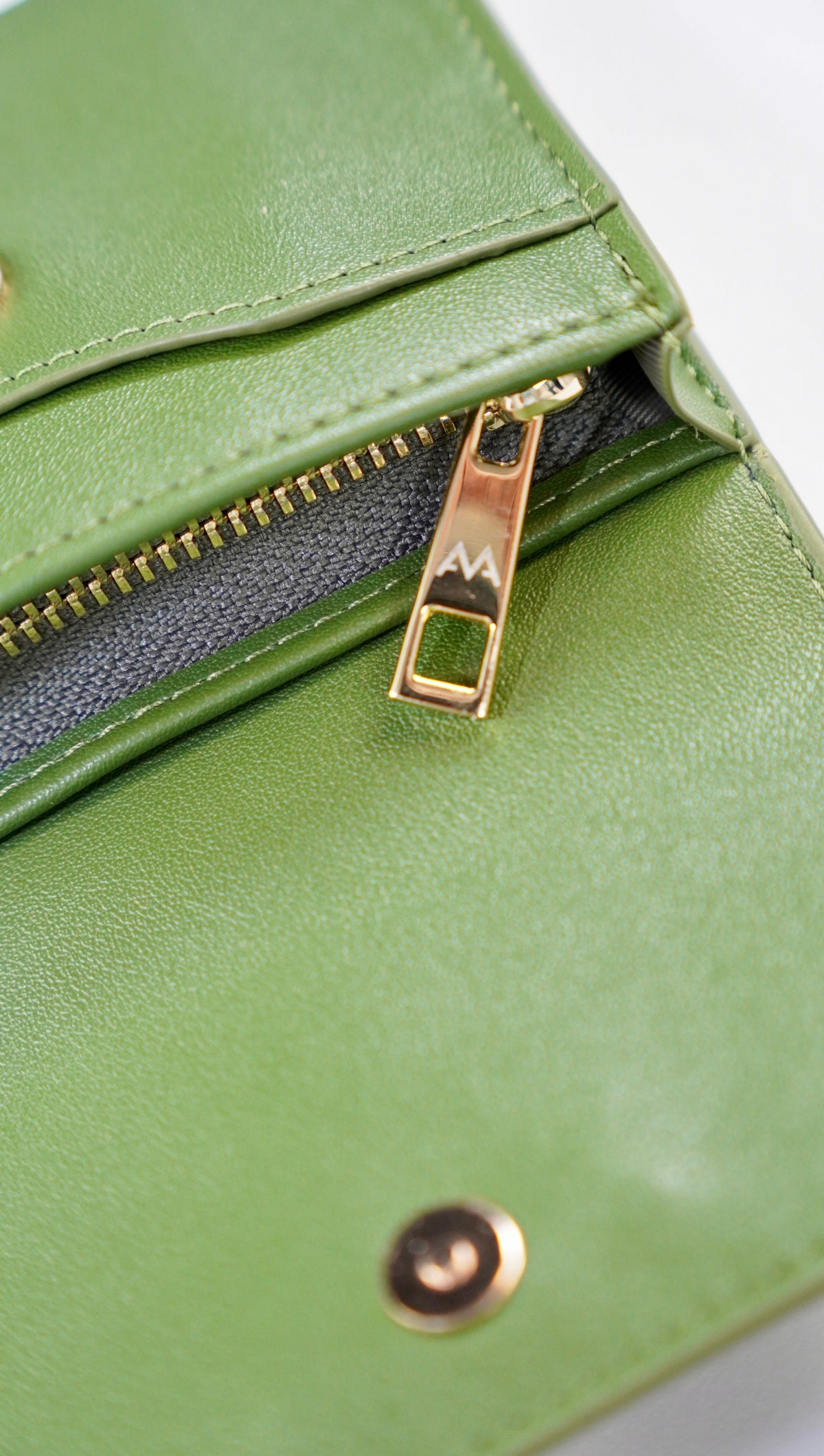 Close-up of a green leather wallet or clutch with a gold zipper pull and a snap button closure, set against a plain white background.