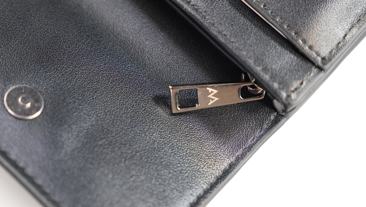 Close-up of a black leather wallet showing a silver metal snap button and a silver zipper pull engraved with a stylized 'M' logo.