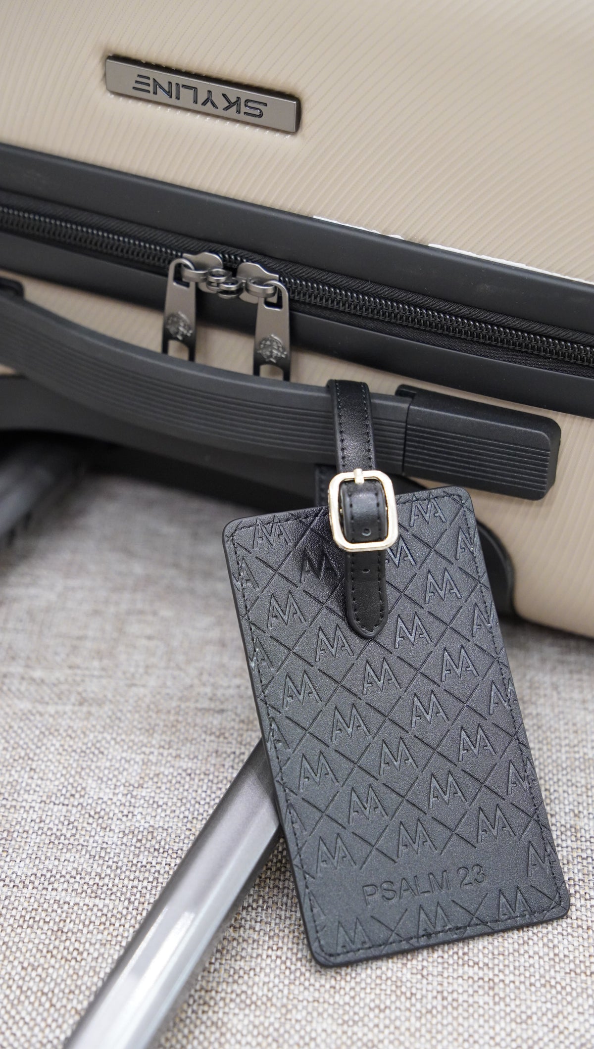 A dark gray embossed leather luggage tag with a gold buckle, attached to a black strap on a beige suitcase.