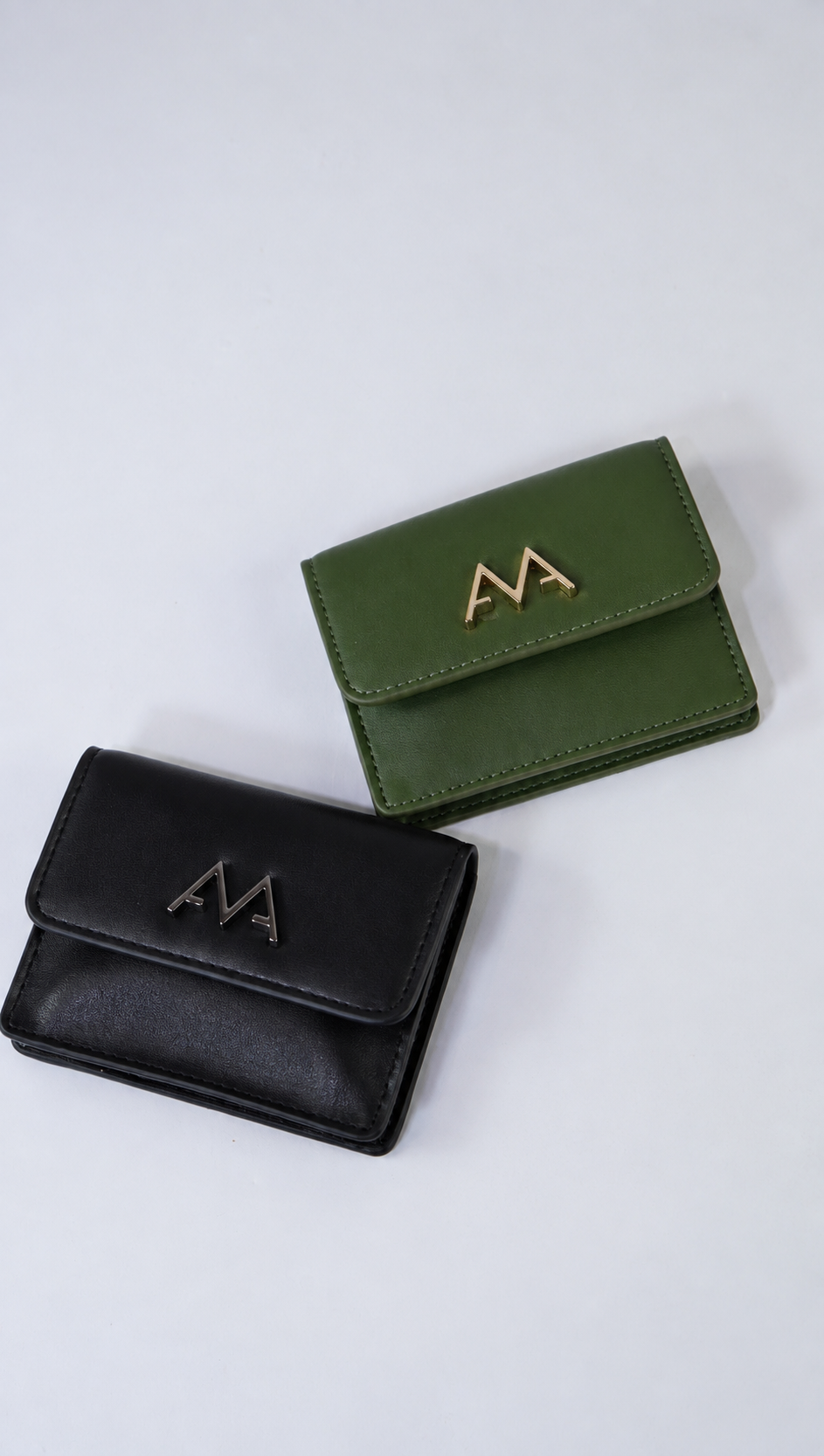 Two small bifold wallets, one black and one olive green, both with a metallic 'M' logo on the front, displayed on a light gray background.