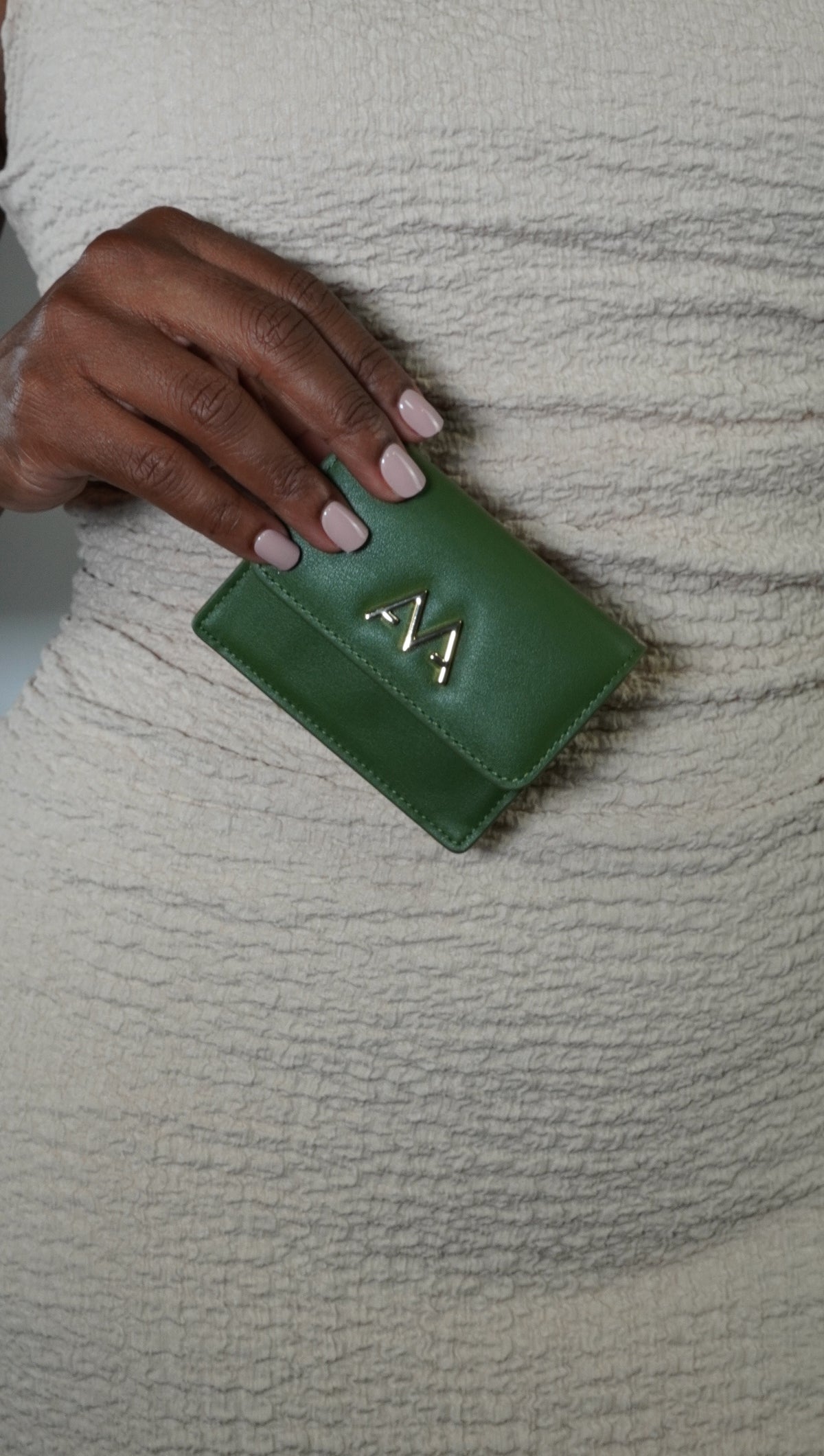 A close-up of a hand holding a small green leather wallet with a gold-tone 'M' logo, set against a textured cream knit top.