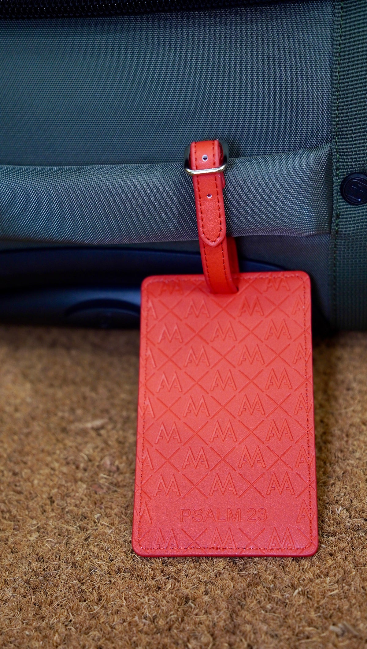 A coral red leather luggage tag with an embossed geometric pattern and the text 'PSALM 23' at the bottom, hanging from a matching leather loop on a dark gray bag over a brown textured surface.