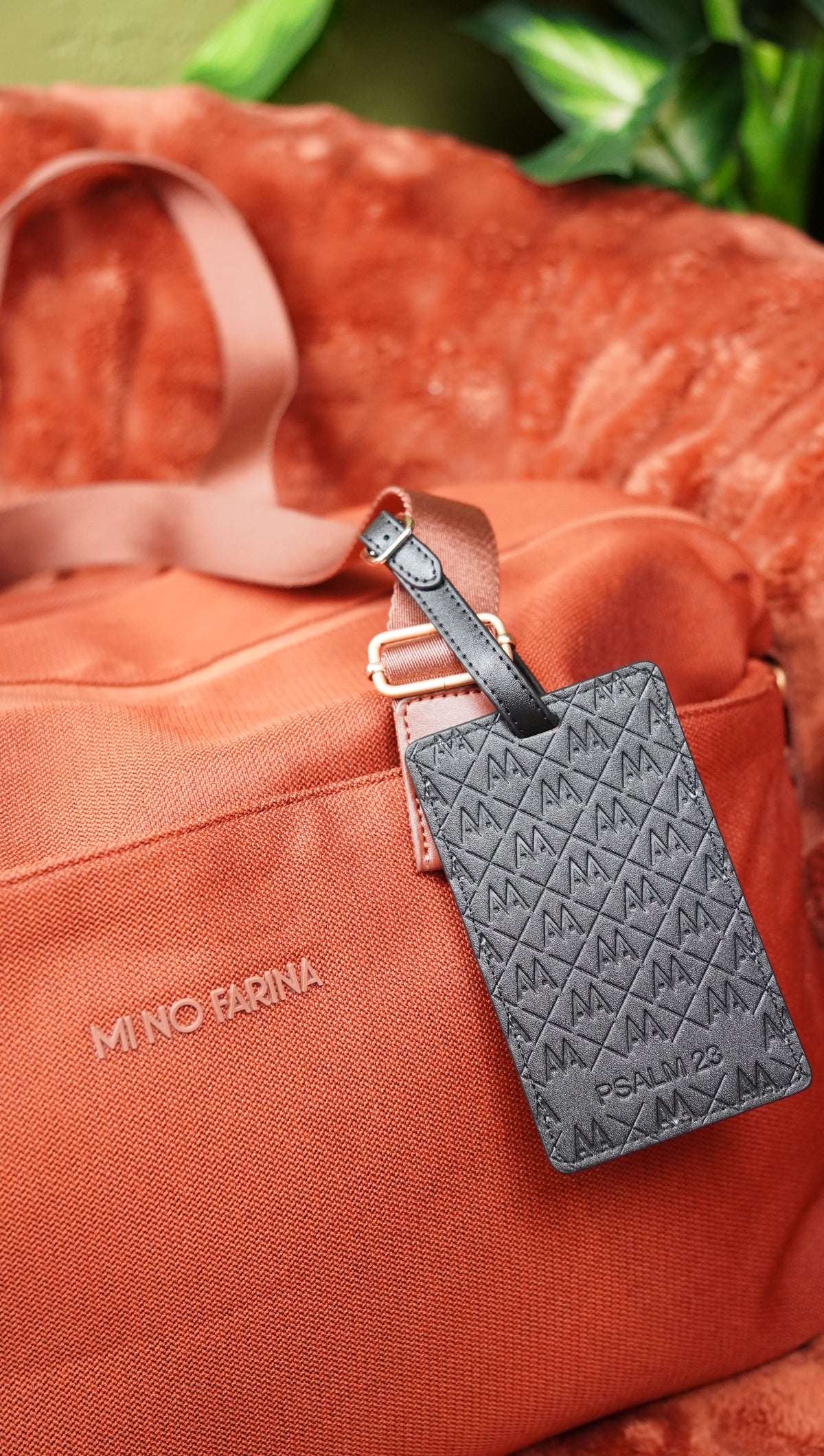An orange canvas duffel bag with a matching orange strap, featuring a dark gray embossed leather luggage tag attached. The brand name 'MINO FARINA' is printed in white on the front of the bag.