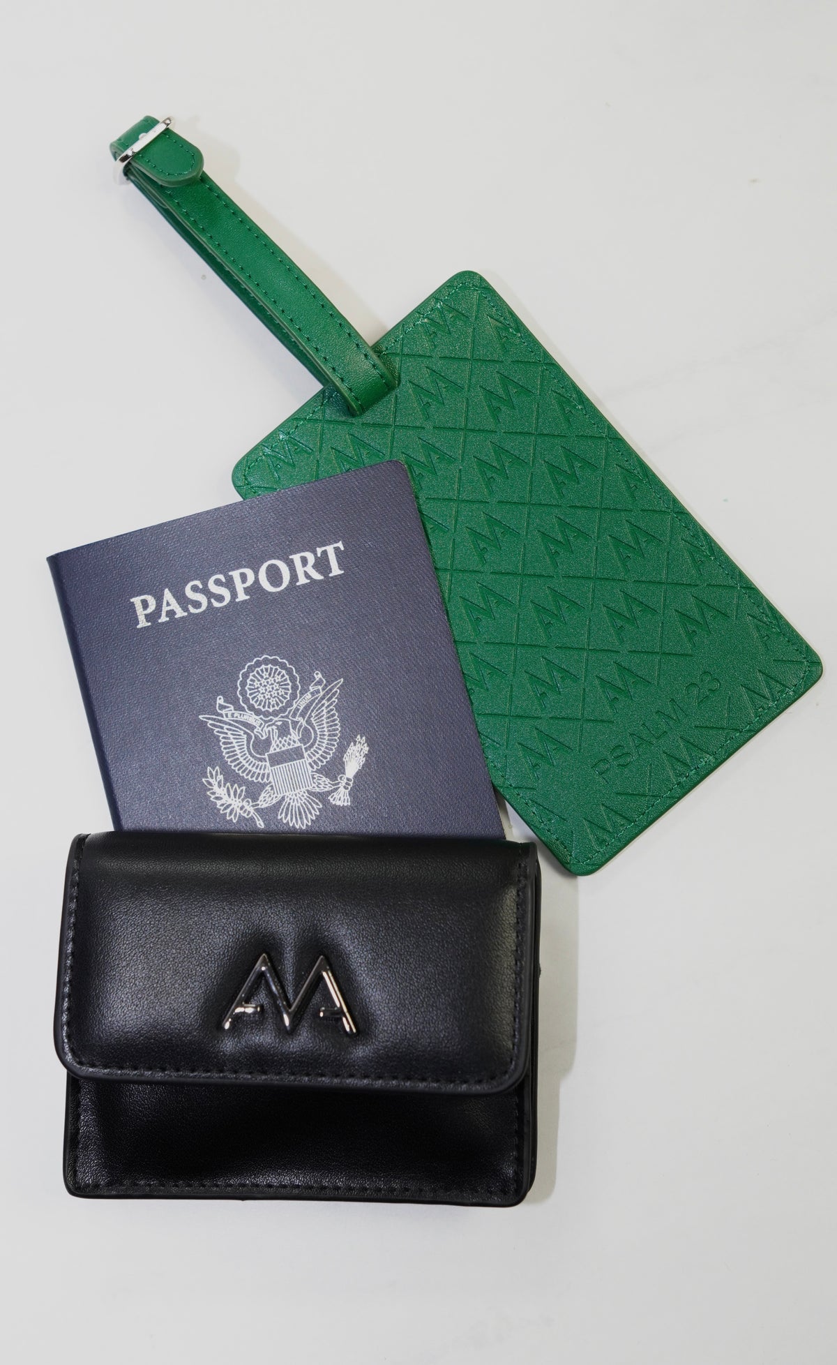 A black leather wallet with a silver 'M' logo, a navy blue passport, and a green embossed luggage tag, arranged on a white background.